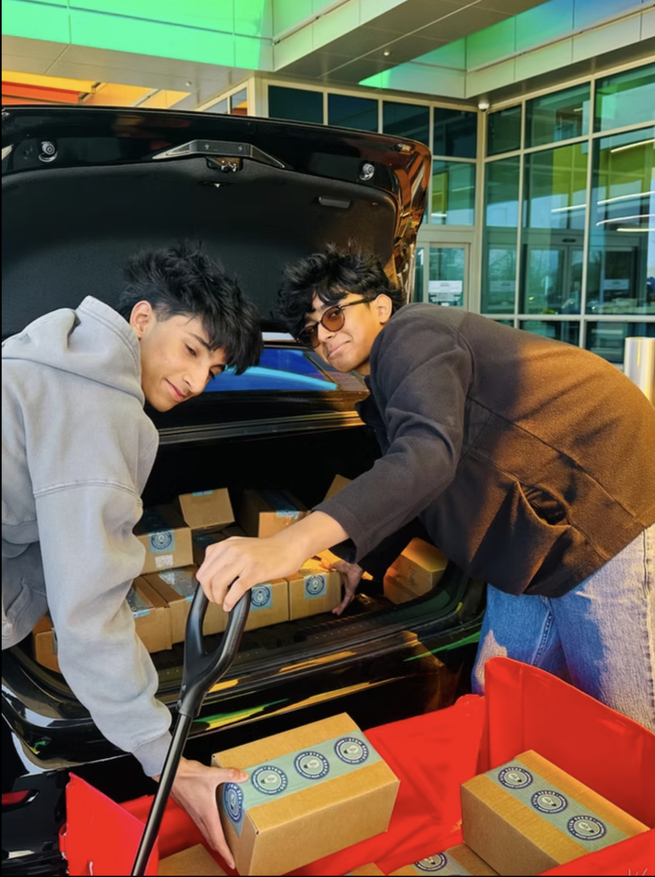 Two students unloading boxed STEM kits from a car trunk