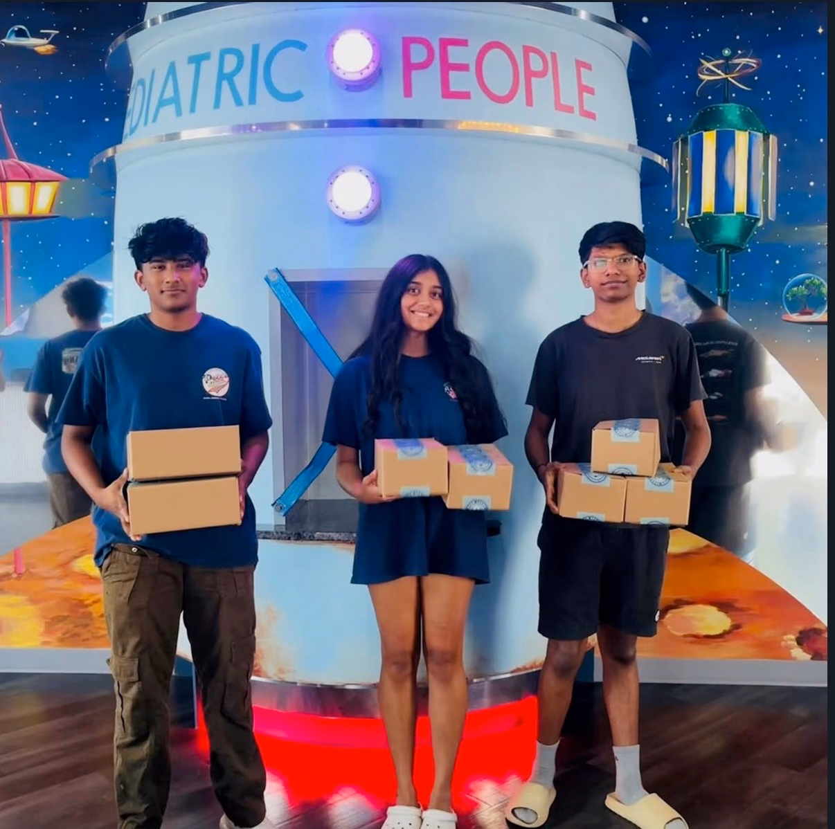 Three chapter members holding STEMKit boxes in front of the Pediatric People rocket ship