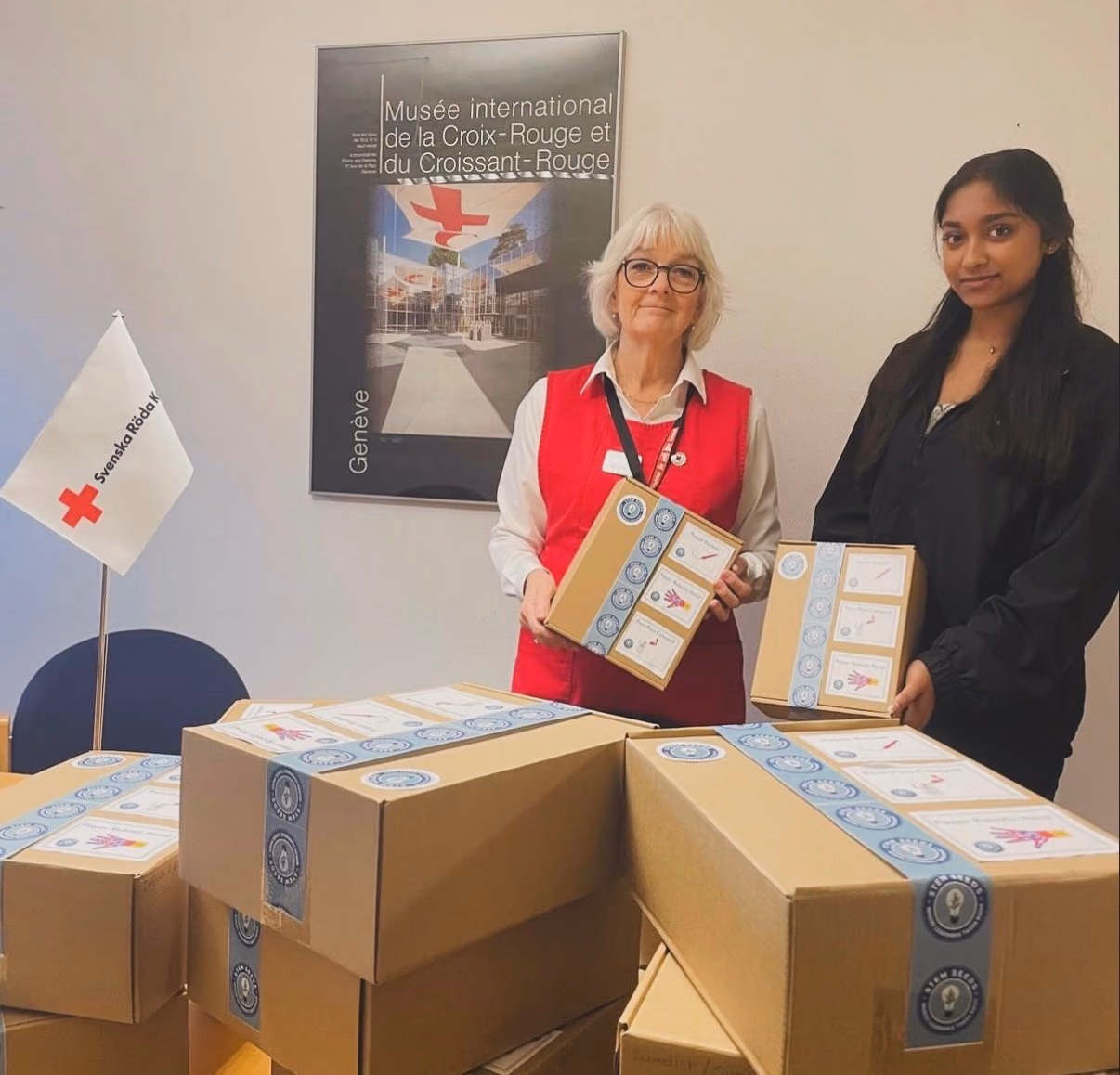Medha Sridar and a Swedish Red Cross volunteer holding STEMKits behind a table of boxes