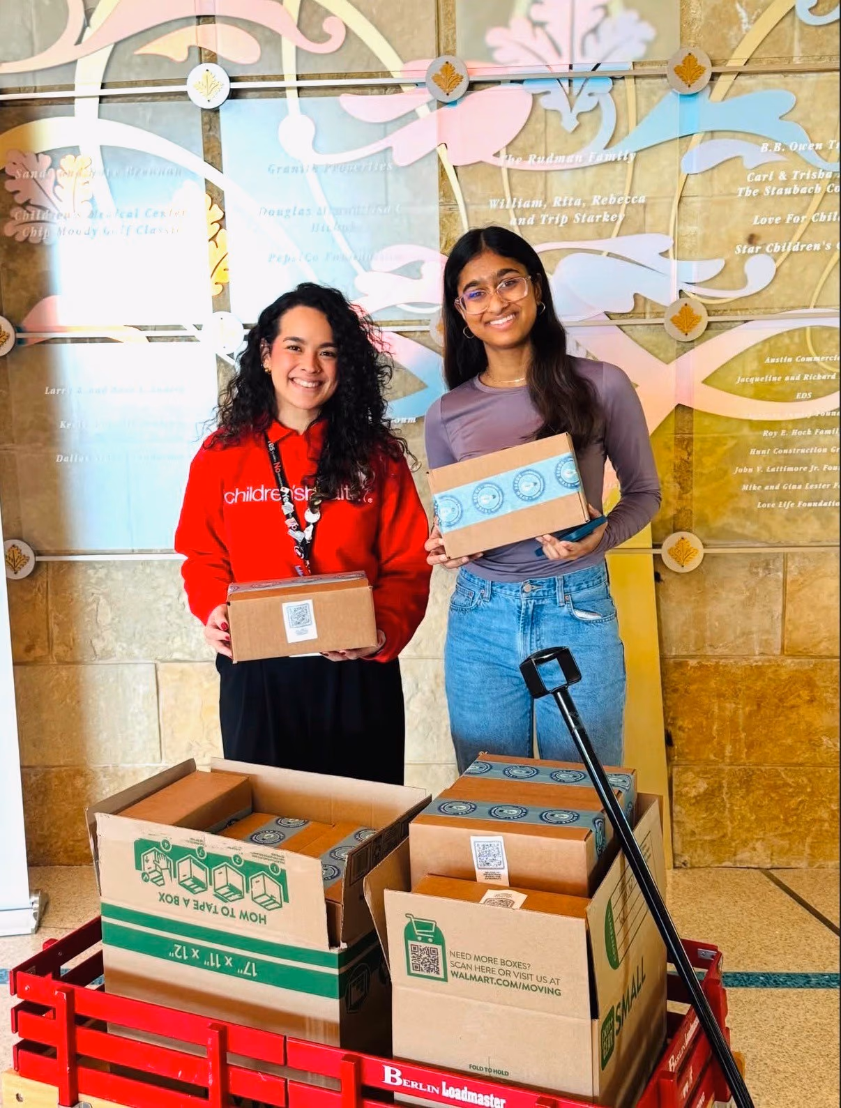 A chapter member and a Children's Health staff member holding STEMKits beside a wagon full of boxes